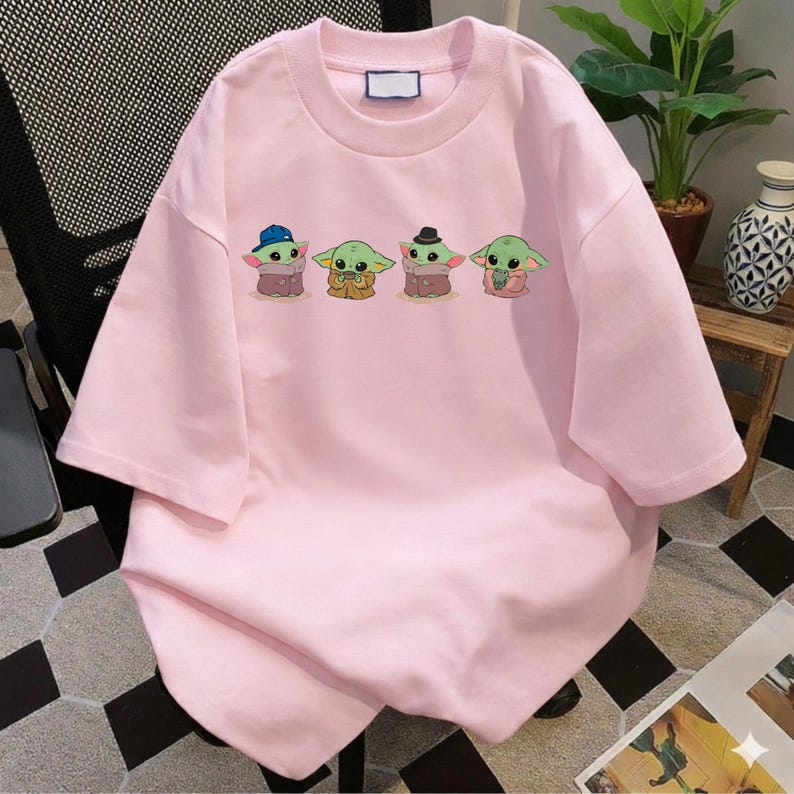 Pu&ograve; includere: Una t-shirt a maniche corte color rosa chiaro con una stampa grafica di quattro piccole creature verdi dalle grandi orecchie. Le creature sono raffigurate in fila, la prima indossa un berretto blu, la terza un cappello nero e la seconda tiene una tazza.