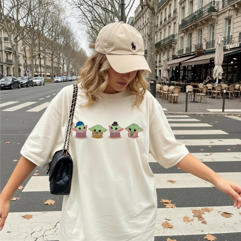 Pu&ograve; includere: Una t-shirt a maniche corte color bianco sporco con una stampa grafica di quattro piccole creature verdi con vari cappelli e abiti, abbinata a un berretto da baseball beige con un piccolo logo scuro e una borsa a tracolla nera trapuntata.