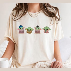 Pu&ograve; includere: Una t-shirt a maniche corte color crema con una stampa grafica di quattro piccole creature verdi. La prima indossa un berretto da baseball blu, la seconda tiene in mano una tazzina, la terza indossa un fedora nero e la quarta tiene in mano una piccola rana verde.