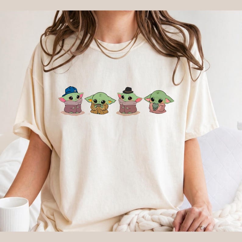 Pu&ograve; includere: Una t-shirt a maniche corte color crema con una stampa grafica di quattro piccole creature verdi. La prima indossa un berretto da baseball blu, la seconda tiene in mano una tazzina, la terza indossa un fedora nero e la quarta tiene in mano una piccola rana verde.