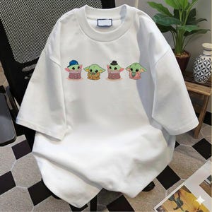 Pu&ograve; includere: Una t-shirt bianca a maniche corte con una stampa grafica di quattro piccole creature verdi dalle grandi orecchie. I personaggi sono raffigurati in fila, il primo indossa un berretto blu, il secondo una tunica marrone, il terzo un cappello scuro e il quarto una barba.