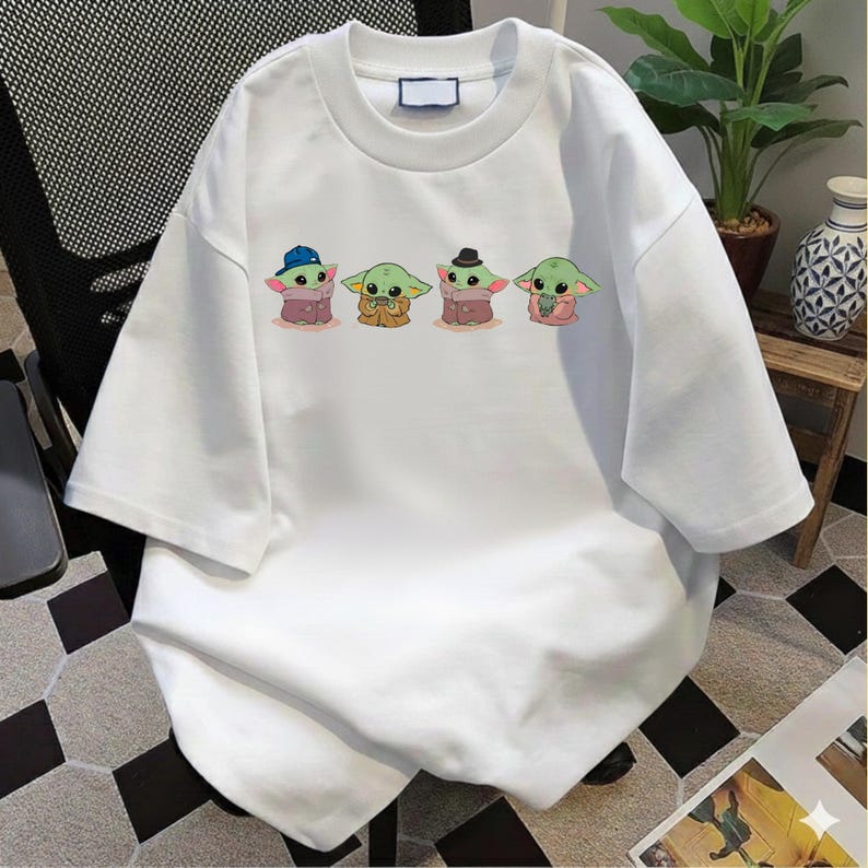 Pu&ograve; includere: Una t-shirt bianca a maniche corte con una stampa grafica di quattro piccole creature verdi dalle grandi orecchie. I personaggi sono raffigurati in fila, il primo indossa un berretto blu, il secondo una tunica marrone, il terzo un cappello scuro e il quarto una barba.