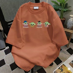 Pu&ograve; includere: Una t-shirt a maniche corte color arancio bruciato con una stampa grafica di quattro piccole creature verdi dalle grandi orecchie. I personaggi sono raffigurati con vari abiti, tra cui un cappello blu, una tunica marrone, un cappello nero e uno scialle rosa.