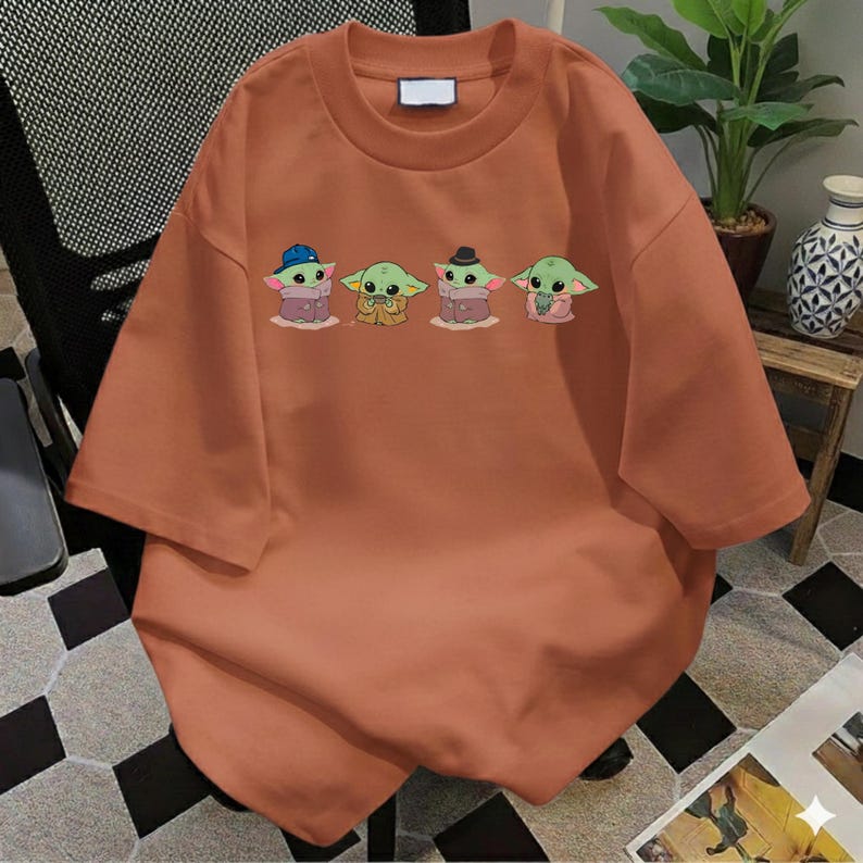 Pu&ograve; includere: Una t-shirt a maniche corte color arancio bruciato con una stampa grafica di quattro piccole creature verdi dalle grandi orecchie. I personaggi sono raffigurati con vari abiti, tra cui un cappello blu, una tunica marrone, un cappello nero e uno scialle rosa.