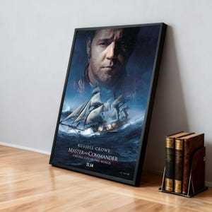 Könnte beinhalten: Ein gerahmtes Filmplakat für "Master and Commander: The Far Side of the World" mit Russell Crowe. Das Poster zeigt ein Schiff auf See mit stürmischem Hintergrund. Das Poster lehnt an einer Wand neben einem Satz Bücher.