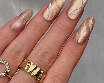 May include: Close-up of a hand with long, almond-shaped nails. The nails are painted with a nude base and gold wave designs. A gold ring with a wavy design is on the ring finger, and a gold ring with a heart-shaped design is on the index finger.