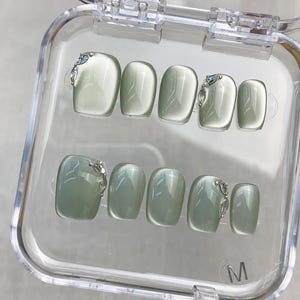 Può includere: Un set di dieci unghie finte color verde salvia con finitura effetto occhio di gatto, disposte in una custodia in plastica trasparente. Alcune unghie presentano piccoli accenti metallici color argento con cristalli chiari. La lettera M è stampata sulla custodia.