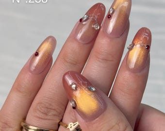May include: Oval-shaped nails with a glossy finish, featuring a warm orange and gold cat-eye effect. The nails are adorned with small clear and dark red rhinestones. Text on the image reads N°:238, Aura Nails, and Ovale Moyen – 21 mm.