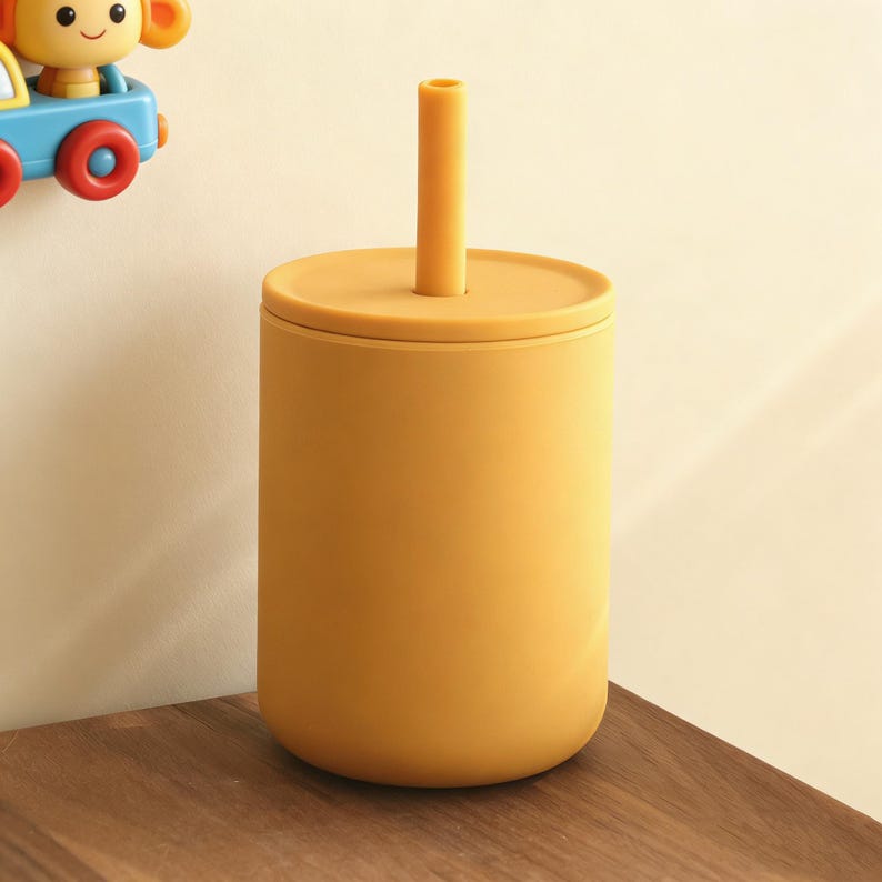 Puede incluir: Vaso de silicona amarillo mostaza con tapa y pajita a juego. El vaso es cil&iacute;ndrico con bordes redondeados. La pajita es recta y se mantiene en posici&oacute;n vertical. El vaso est&aacute; sobre una superficie de madera.
