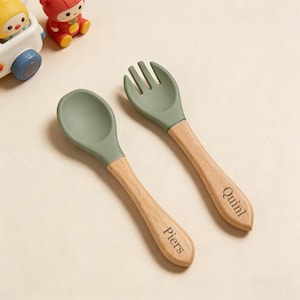 Puede incluir: Un juego de cuchara y tenedor de silicona verde con mangos de madera. La cuchara y el tenedor están diseñados para bebés y niños pequeños. Los mangos de madera están grabados con los nombres "Piers" y "Quinl".