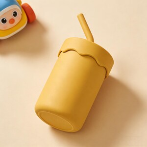 May include: Mustard yellow cup with a matching straw. The cup has a wavy rim and a cylindrical shape. A yellow and blue toy is in the background.