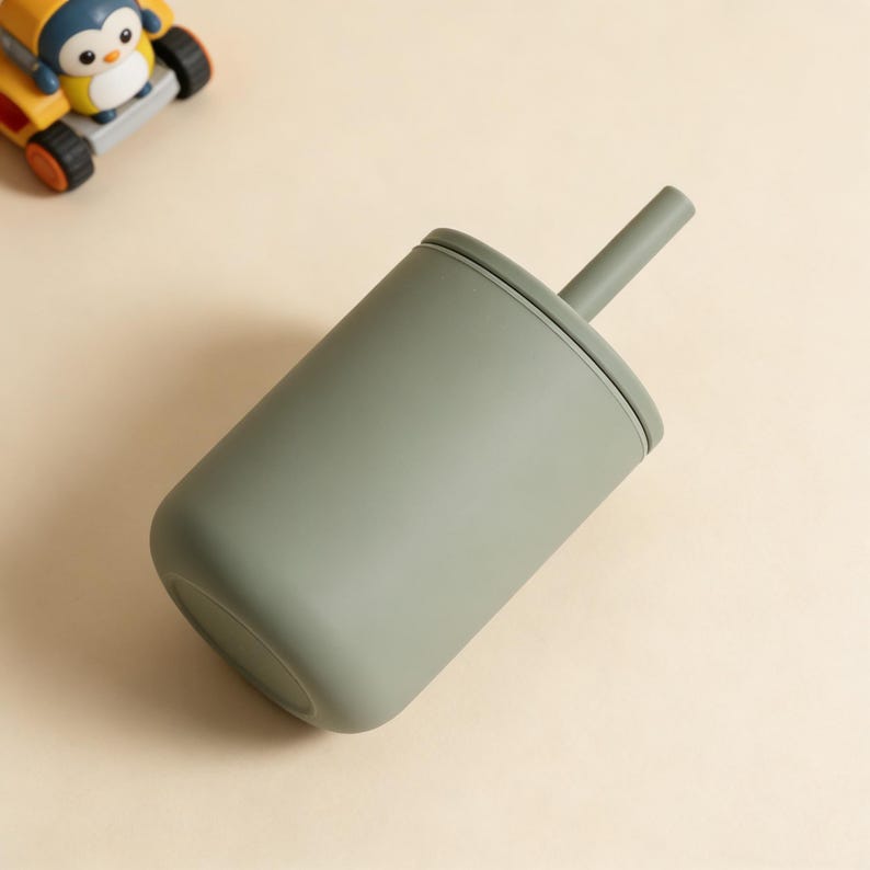 Puede incluir: Una taza de silicona verde salvia con tapa y pajita a juego. La taza est&aacute; en &aacute;ngulo, mostrando su base redondeada y su superficie lisa. Un coche de juguete amarillo y negro con un ping&uuml;ino est&aacute; en el fondo.