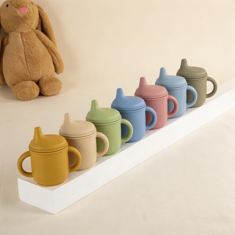 May include: A row of colorful silicone sippy cups in shades of yellow, beige, green, blue, pink, and olive green. Each cup has a handle and a lid with a spout. A plush bunny is in the background.
