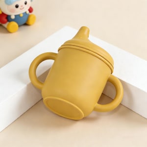 May include: Mustard yellow baby sippy cup with two handles and a spout. The cup is made of a soft, flexible material and has a rounded base. A small toy is visible in the background.