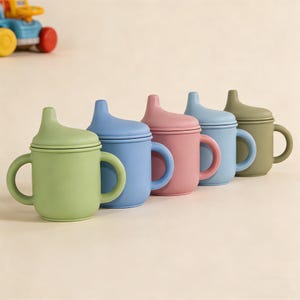 May include: A row of colorful baby sippy cups in shades of green, blue, pink, and olive green. Each cup has a matching lid and two handles. The cups are made of a soft, matte material.