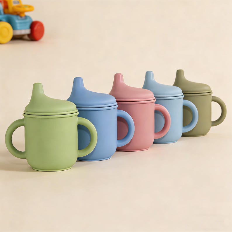 May include: A row of colorful baby sippy cups in shades of green, blue, pink, and olive green. Each cup has a matching lid and two handles. The cups are made of a soft, matte material.