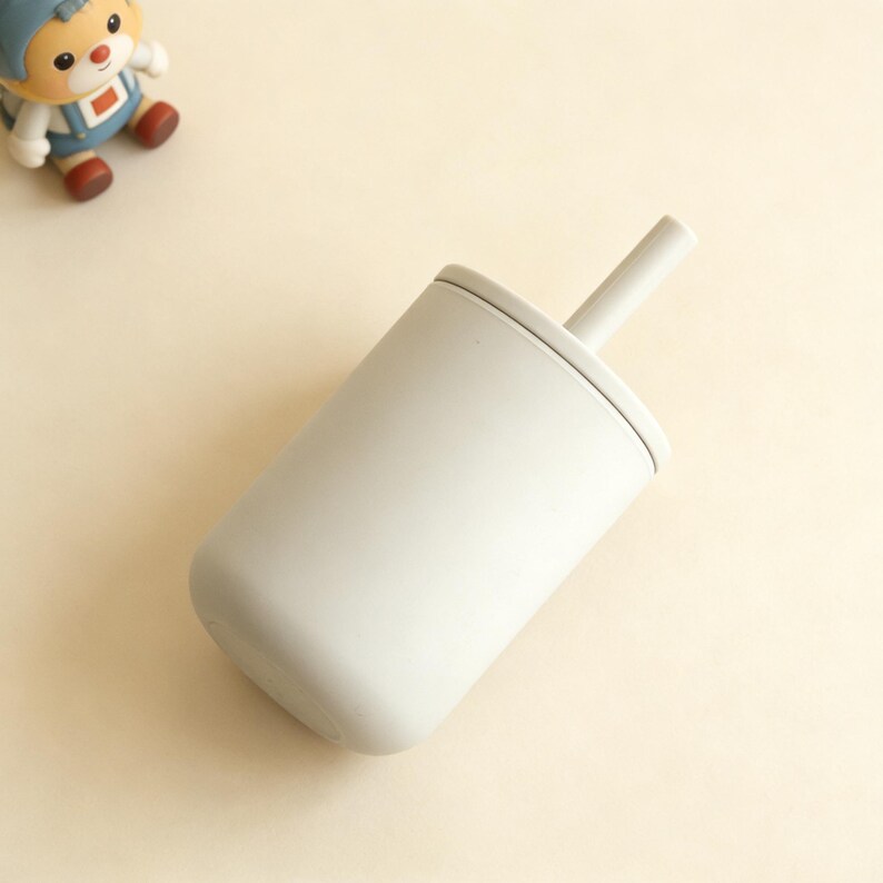 Puede incluir: Un vaso para sorber de color gris claro con tapa y pajita a juego. La taza es de material mate y tiene una base redondeada. Un peque&ntilde;o juguete es visible en el fondo.