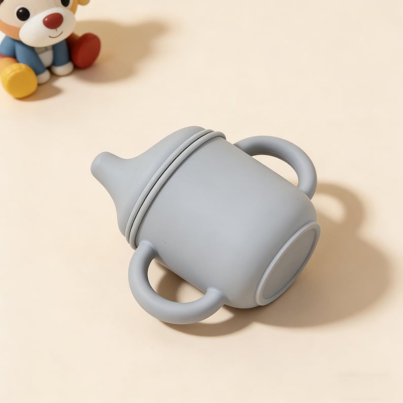 May include: A gray silicone baby bottle with two handles and a nipple. The bottle is designed for easy gripping and feeding. A small toy is visible in the background.