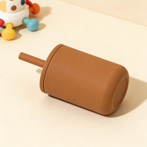 Puede incluir: Una taza de silicona marr&oacute;n con una pajita a juego. La taza es cil&iacute;ndrica con bordes redondeados. Un juguete colorido es parcialmente visible en el fondo. La taza est&aacute; dise&ntilde;ada para beb&eacute;s y ni&ntilde;os peque&ntilde;os.