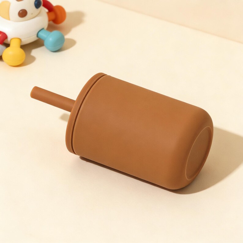 Puede incluir: Una taza de silicona marr&oacute;n con una pajita a juego. La taza es cil&iacute;ndrica con bordes redondeados. Un juguete colorido es parcialmente visible en el fondo. La taza est&aacute; dise&ntilde;ada para beb&eacute;s y ni&ntilde;os peque&ntilde;os.