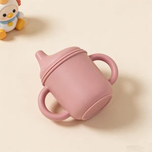 May include: A dusty rose-colored sippy cup with two handles. The cup has a rounded base and a spout with a lid. The cup is made of a soft, flexible material. A toy chicken is in the background.