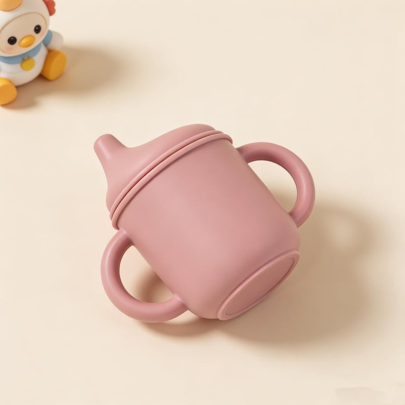 May include: A dusty rose-colored sippy cup with two handles. The cup has a rounded base and a spout with a lid. The cup is made of a soft, flexible material. A toy chicken is in the background.