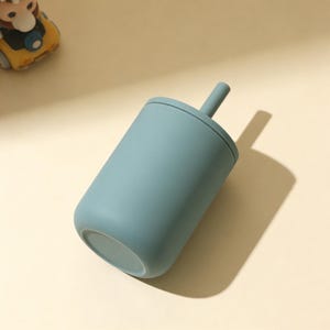 Puede incluir: Una taza de color verde azulado con tapa y pajita a juego. La taza tiene una base redondeada y un cuerpo cil&iacute;ndrico. La pajita se extiende desde el centro de la tapa. La taza est&aacute; hecha de un material mate. Un coche de juguete es visible en el fondo.