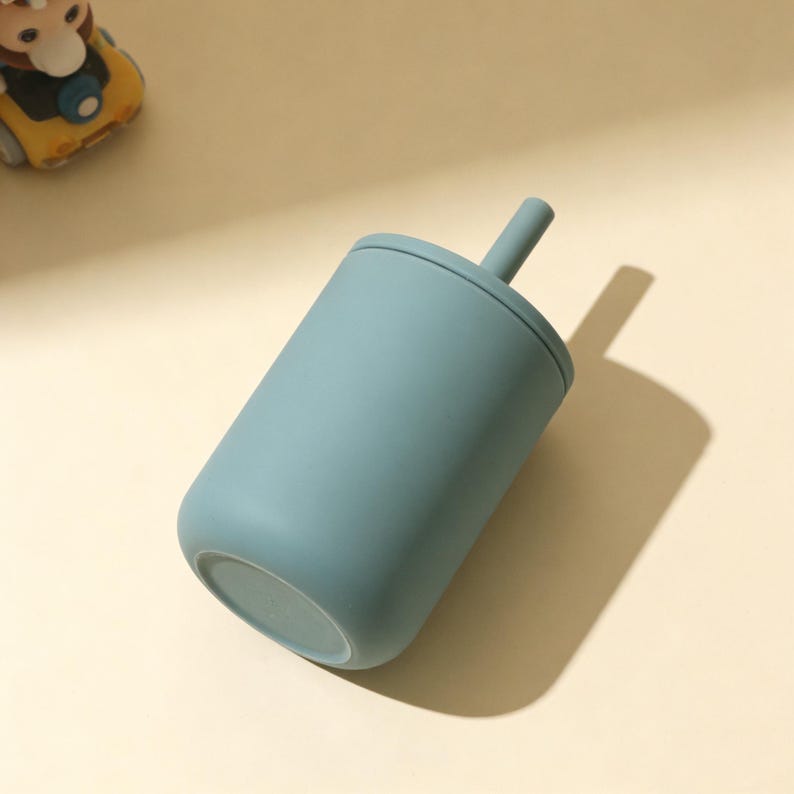 Puede incluir: Una taza de color verde azulado con tapa y pajita a juego. La taza tiene una base redondeada y un cuerpo cil&iacute;ndrico. La pajita se extiende desde el centro de la tapa. La taza est&aacute; hecha de un material mate. Un coche de juguete es visible en el fondo.