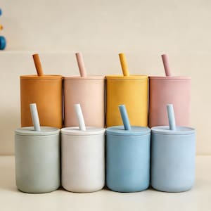Puede incluir: Ocho tazas de silicona de colores con tapas y pajitas. Las tazas son en tonos naranja, rosa, amarillo y azul. Un juguete est&aacute; en el fondo.