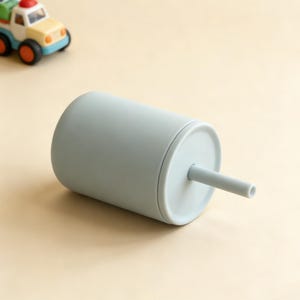Puede incluir: Un vaso para sorber de color azul claro con tapa y pajita a juego. El vaso es cil&iacute;ndrico y se apoya sobre una superficie de color beige claro. Un peque&ntilde;o cami&oacute;n de juguete est&aacute; en el fondo.