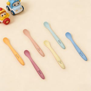 May include: Six pastel-coloured baby spoons, each with a name printed on the handle. The spoons are in shades of yellow, orange, pink, purple, light blue, and blue. Two toy cars are in the top left corner.