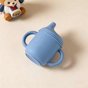 May include: A light blue baby sippy cup with two handles and a spout. The cup is made of a soft, flexible material. A cartoon bear figure is in the background.