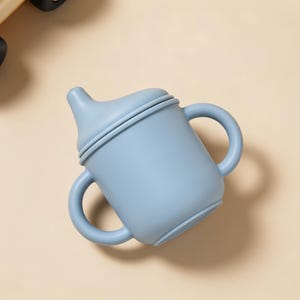 May include: A light blue baby sippy cup with two handles and a spout. The cup is made of a soft, matte material. A yellow and cream-colored toy car is partially visible in the upper left corner.