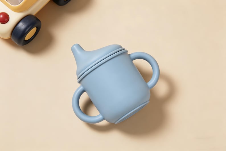 May include: A light blue baby sippy cup with two handles and a spout. The cup is made of a soft, matte material. A yellow and cream-colored toy car is partially visible in the upper left corner.