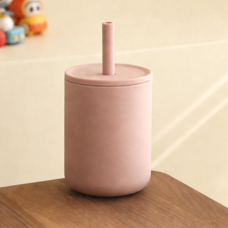 Puede incluir: Una taza de silicona de color rosa polvoriento con tapa y pajita a juego. La taza es cil&iacute;ndrica con bordes redondeados. La pajita es recta y se extiende hacia arriba desde el centro de la tapa. La taza est&aacute; sobre una superficie de madera.