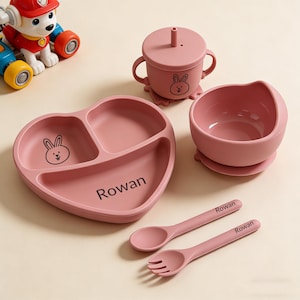 May include: A pink baby feeding set includes a heart-shaped divided plate, a bowl, a sippy cup, and two utensils. Each piece has a bunny graphic and the name "Rowan".