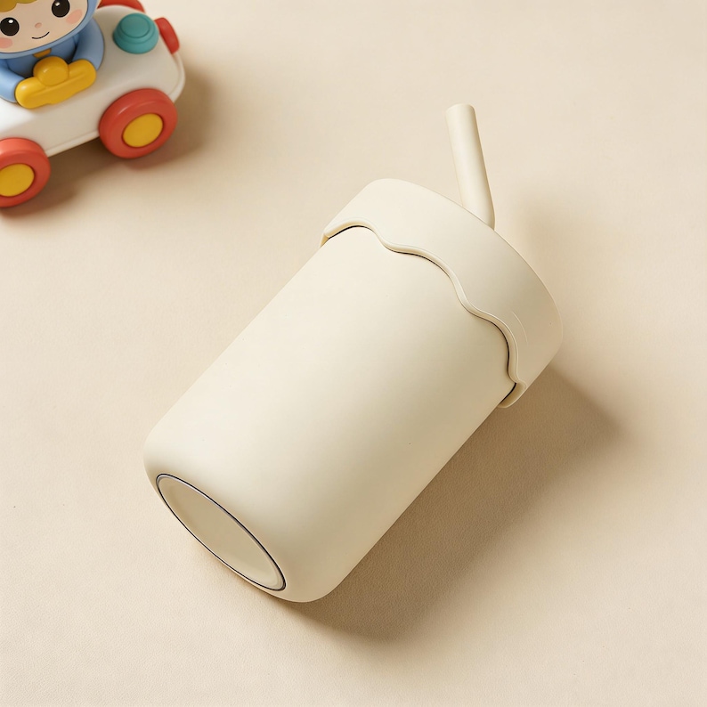 May include: A cream-colored tumbler with a matching lid and straw. The lid has a wavy edge design. A toy car with a blue driver and red and yellow wheels is in the background.