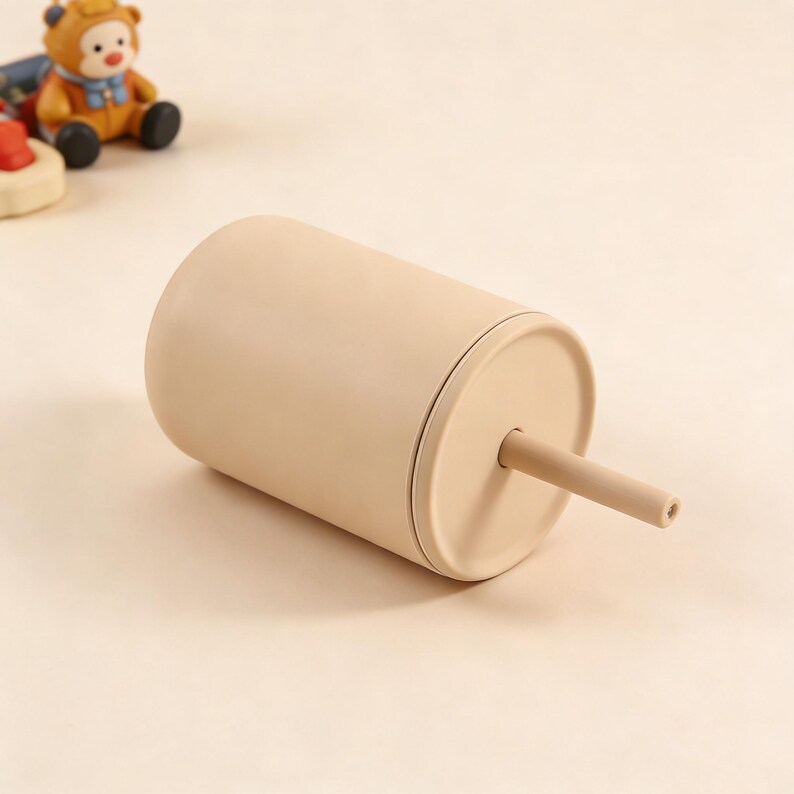 Puede incluir: Un vaso beige con tapa y pajita a juego. El vaso cil&iacute;ndrico parece estar hecho de un material mate. La pajita se extiende desde la tapa en &aacute;ngulo. Un oso de juguete est&aacute; en el fondo.