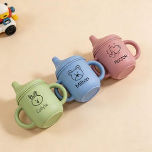 May include: Three colorful baby sippy cups in green, blue, and pink. Each cup has a handle and a lid with a spout. The cups are decorated with a bunny, bear, and heart design, and the names "Calvin", "Milton", and "Hector" are printed on them.