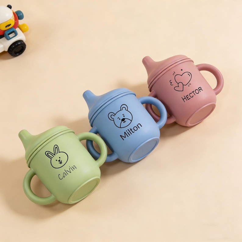May include: Three colorful baby sippy cups in green, blue, and pink. Each cup has a handle and a lid with a spout. The cups are decorated with a bunny, bear, and heart design, and the names "Calvin", "Milton", and "Hector" are printed on them.