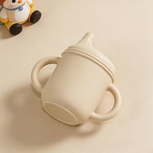 May include: A beige sippy cup with two handles and a spout. The cup is made of a soft, flexible material. A small toy is visible in the background.