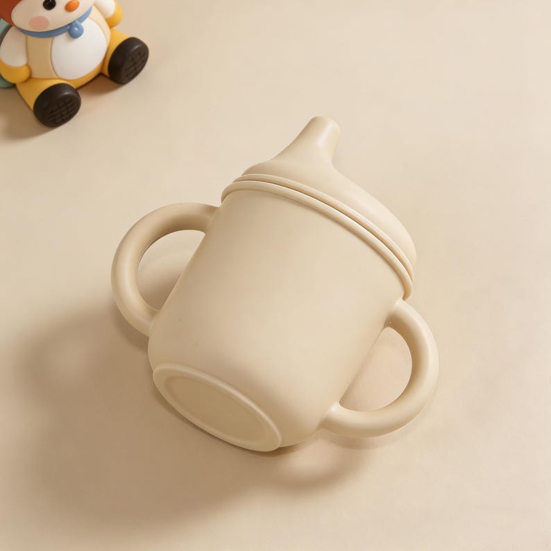 May include: A beige sippy cup with two handles and a spout. The cup is made of a soft, flexible material. A small toy is visible in the background.