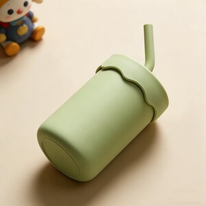 May include: A light green cup with a matching lid and straw. The lid has a wavy edge. The cup is cylindrical and appears to be made of a soft, matte material. A small toy is in the background.