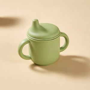 May include: A light green baby sippy cup with a lid and two handles. The cup is made of a soft material and is designed for easy gripping. The sippy cup is on a light tan surface.