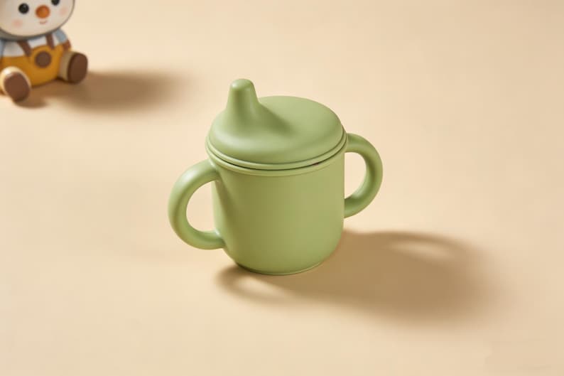 May include: A light green baby sippy cup with a lid and two handles. The cup is made of a soft material and is designed for easy gripping. The sippy cup is on a light tan surface.