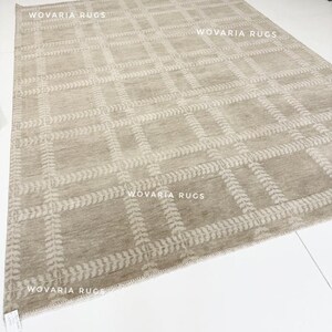 May include: A large, rectangular rug in a neutral beige hue, featuring a grid design. The grid is accented with a lighter beige leaf pattern. The text "WOVARIA RUGS" is visible on the rug.
