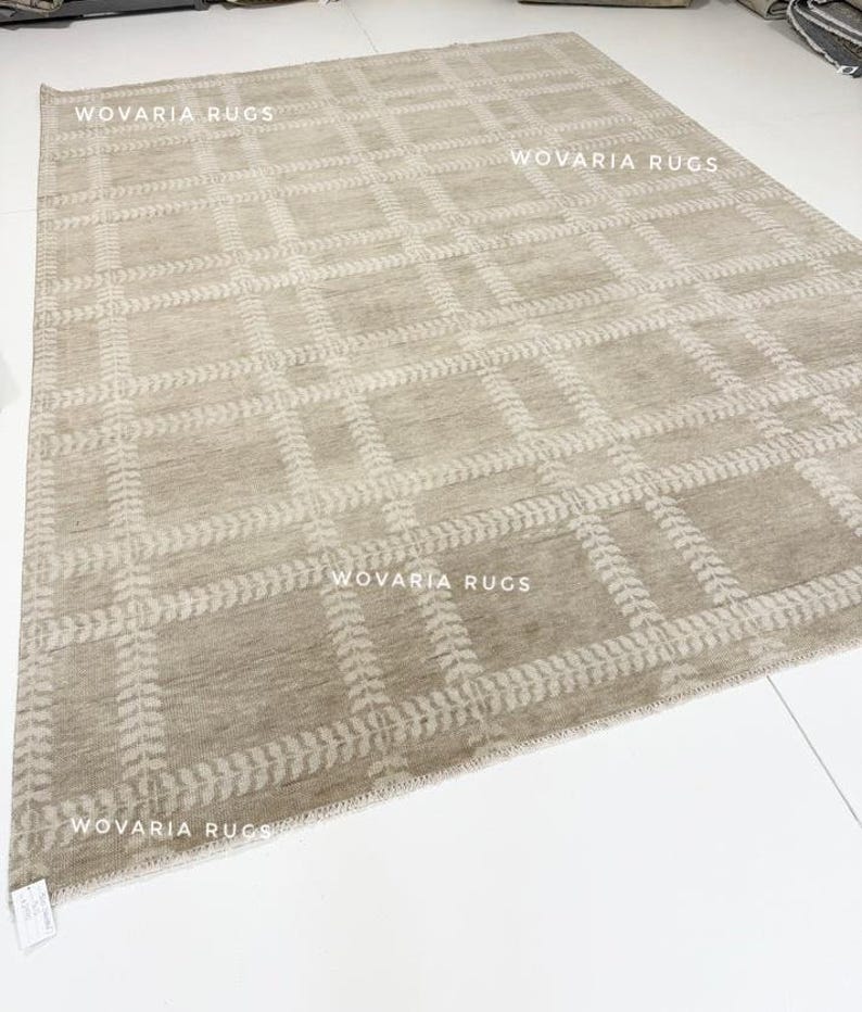 May include: A large, rectangular rug in a neutral beige hue, featuring a grid design. The grid is accented with a lighter beige leaf pattern. The text "WOVARIA RUGS" is visible on the rug.