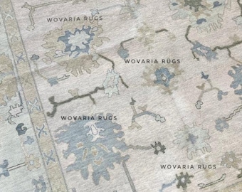 May include: Close-up of a beige rug with a floral pattern in shades of blue, green, and gray. The rug has a distressed look and the text "WOVARIA RUGS" is printed across the surface.