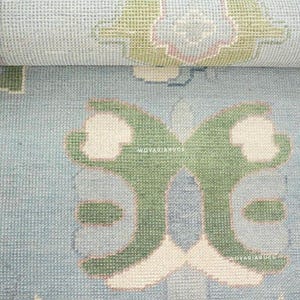 May include: A light blue area rug featuring a repeating pattern of stylized green and cream floral motifs. The rug has a textured, woven surface with the text WOVARIARUGS visible in small white letters across the central design elements.