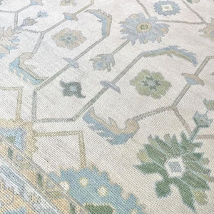 May include: Close-up of a cream-colored rug with a geometric floral pattern. The design features light blue, green, and tan floral and geometric shapes. The rug has a textured appearance.